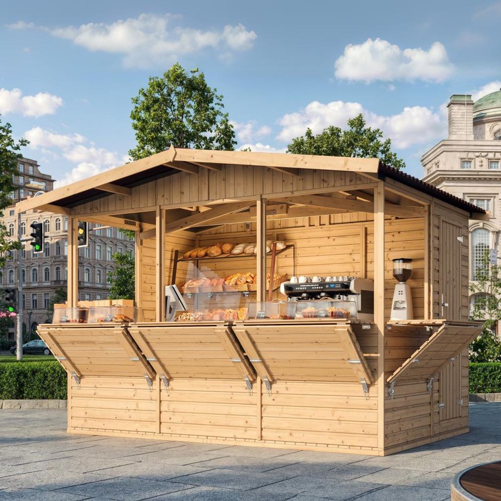 A wooden market stall selling baked goods and coffee, set up on a paved plaza with buildings and trees in the background.