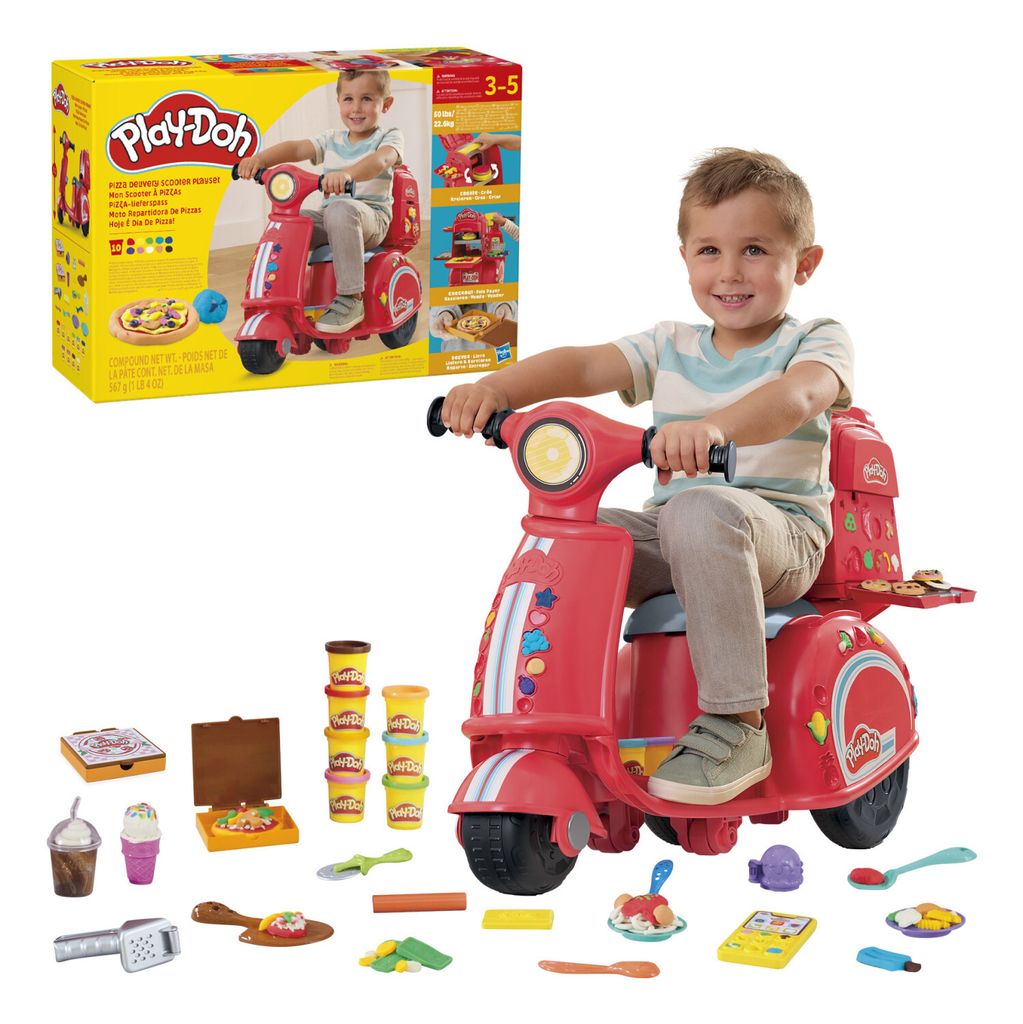 A young boy is riding a red Play-Doh pizza delivery scooter. Various Play-Doh accessories are scattered around the scooter.