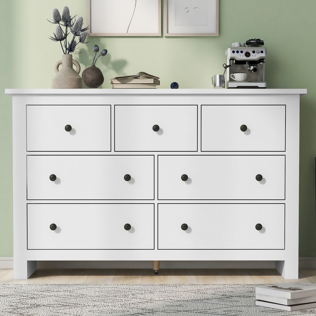 A white wooden dresser with seven drawers is shown in a room with a light green wall and a rug on the floor. Decorative items are placed on top of the dresser.