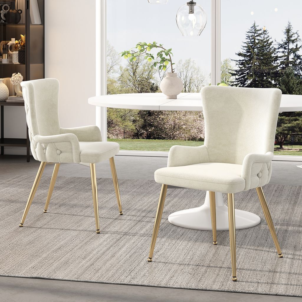Two beige upholstered dining chairs with gold legs are placed in a dining room setting with a white oval table and a large window overlooking a garden.