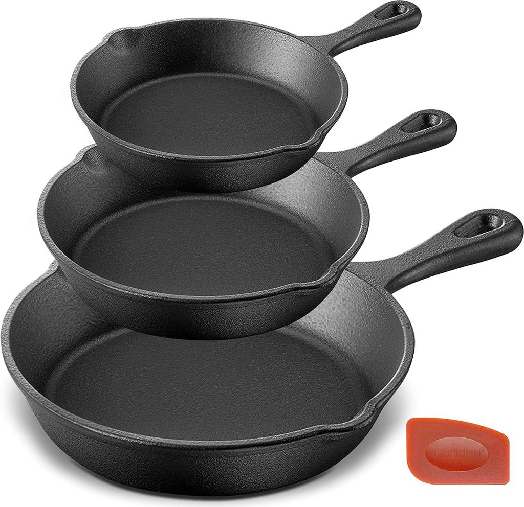 A stack of three black cast iron skillets of varying sizes, with a small orange silicone scraper in the bottom right corner.