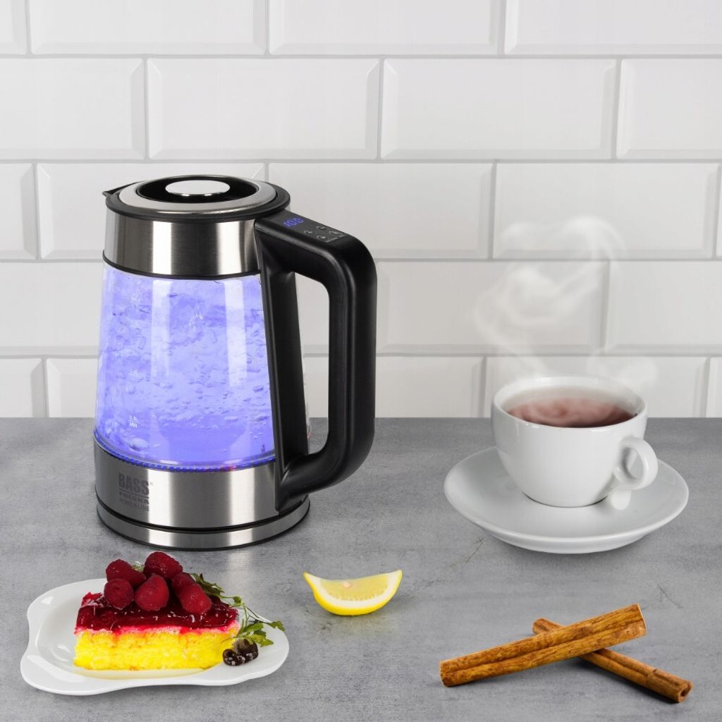 A glass electric kettle with blue light, a cup of tea, a slice of cake, lemon, and cinnamon sticks on a countertop.
