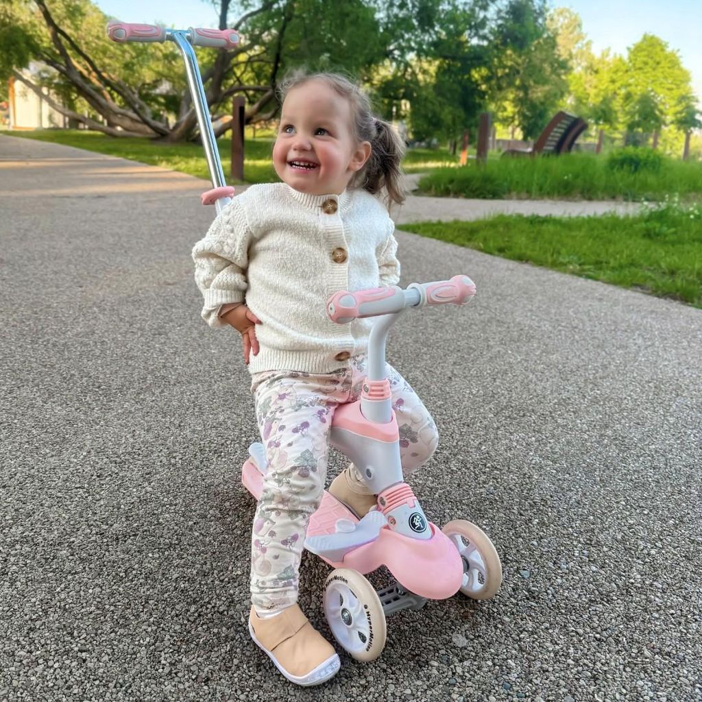 A young child is happily sitting on a pink and white balance scooter in a park.