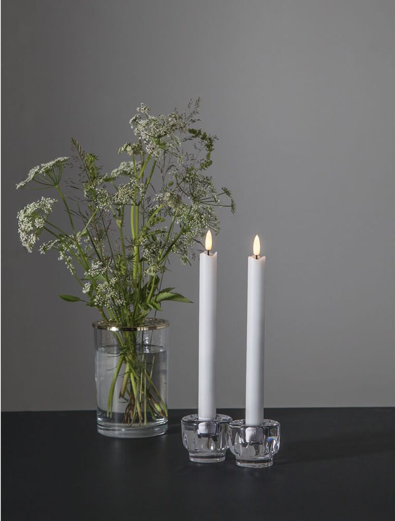 Two white LED candles in crystal candle holders are placed next to a glass vase filled with white flowers on a dark surface.
