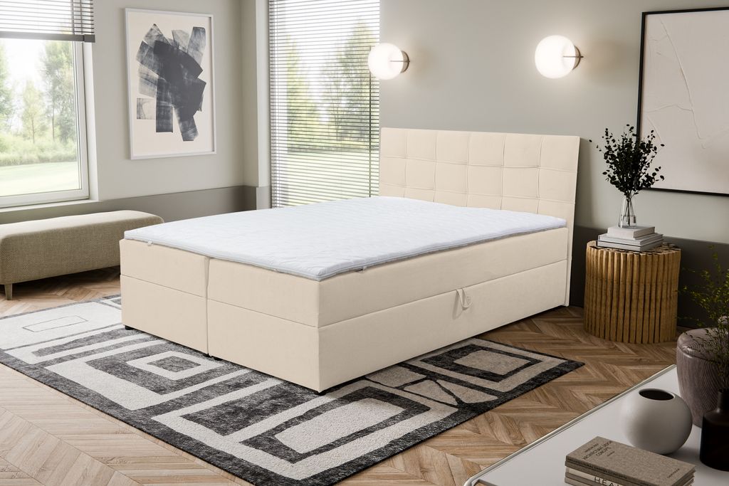 A bedroom scene featuring a beige boxspring bed with storage drawers, a patterned rug, and decorative elements.