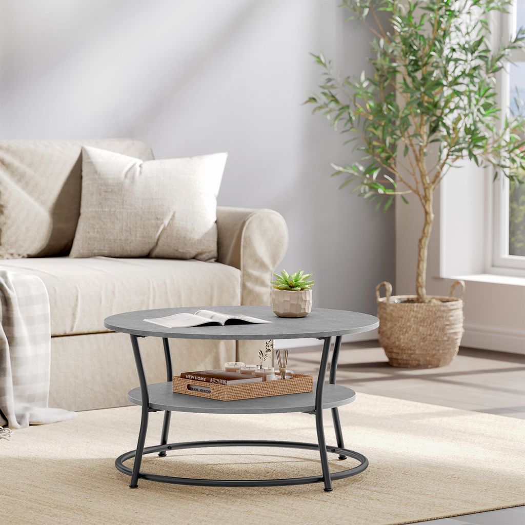 A modern coffee table with a two-tiered design is placed on a rug in front of a sofa. A small plant sits on the top tier, and a tray with items is on the lower tier. A large potted plant is to the right.