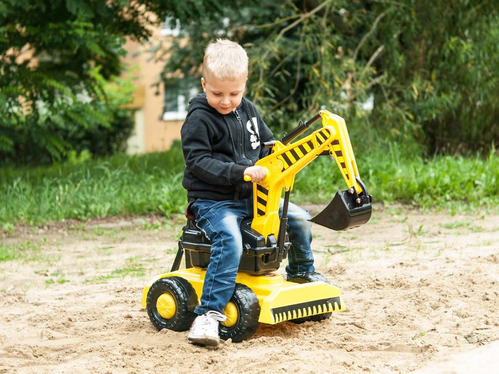 A young boy is playing with a yellow ride-on toy excavator in a sandy area with trees and a building in the background.