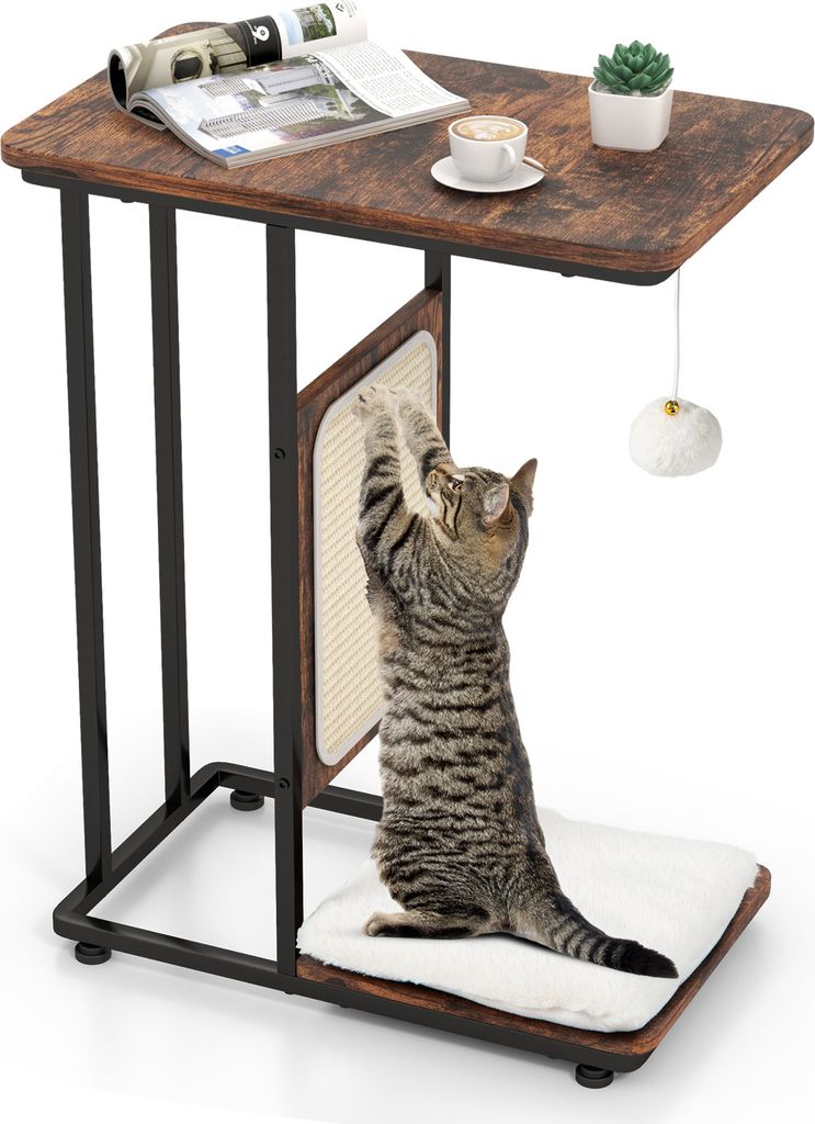 A cat is scratching a sisal board attached to a side table. The table has a wooden top with a magazine, coffee cup, and a small plant on it. A fluffy toy hangs from the table.