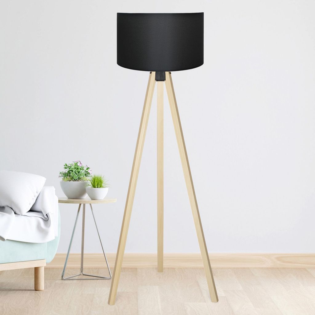 A floor lamp with a black shade and wooden tripod legs is shown in a room with a sofa and a small table with plants.
