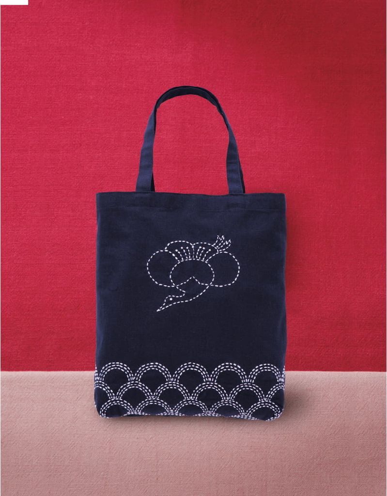 A dark blue tote bag with white embroidery is displayed against a red background and a beige surface.