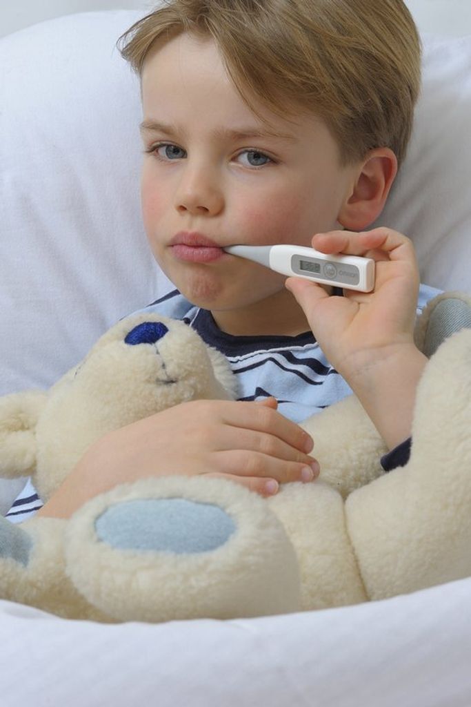 A young boy is lying in bed, holding a teddy bear and taking his temperature with a digital thermometer.