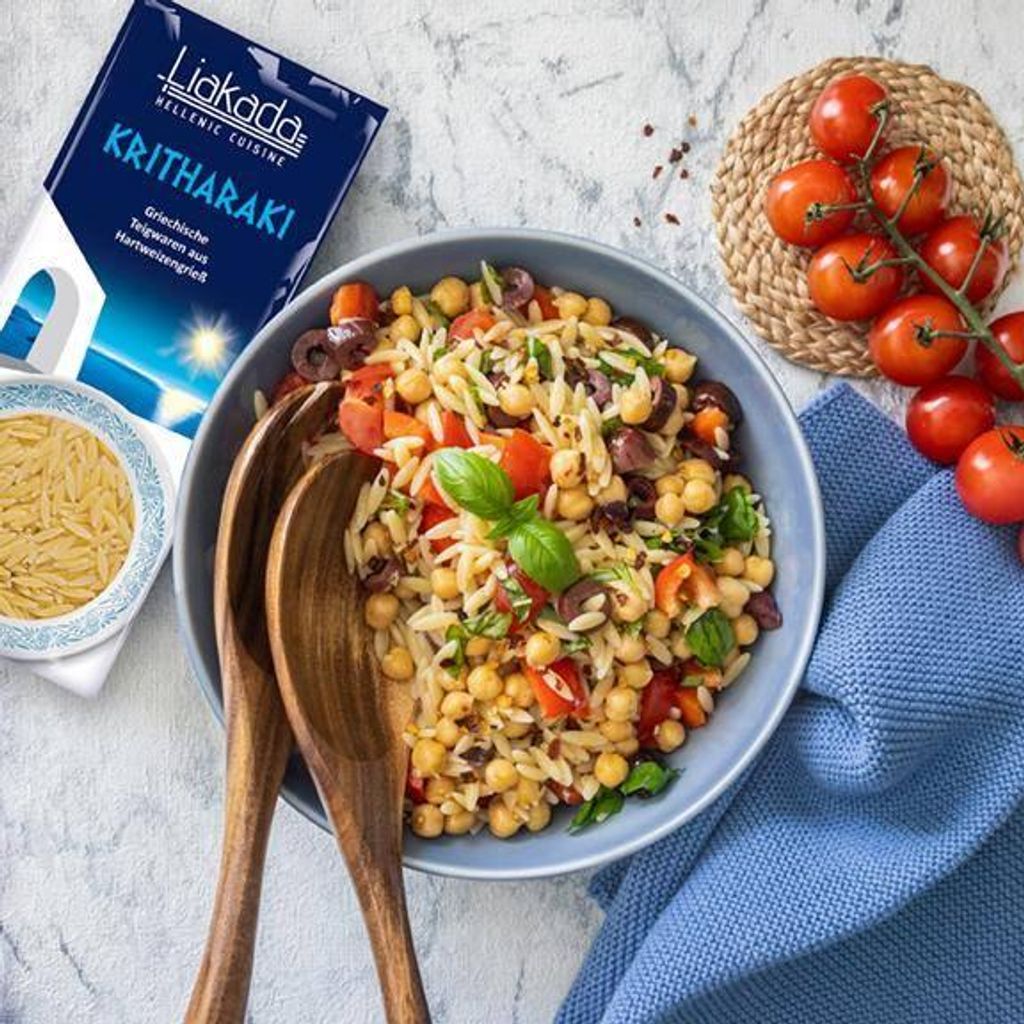 A bowl of orzo pasta salad with chickpeas, tomatoes, and olives, next to a package of Kritharaki pasta and a bunch of cherry tomatoes.
