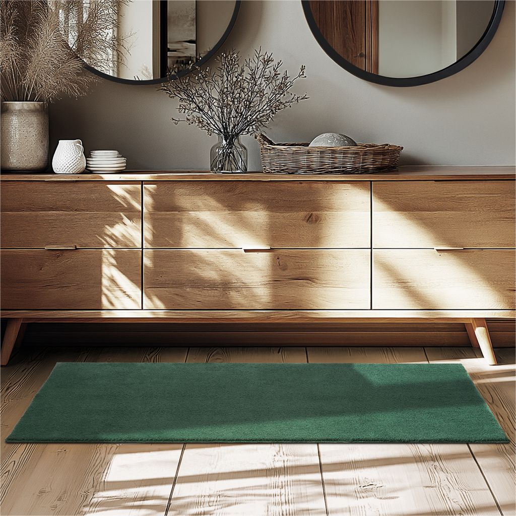 A green rug is placed on a wooden floor in front of a wooden dresser with decorative items on top.