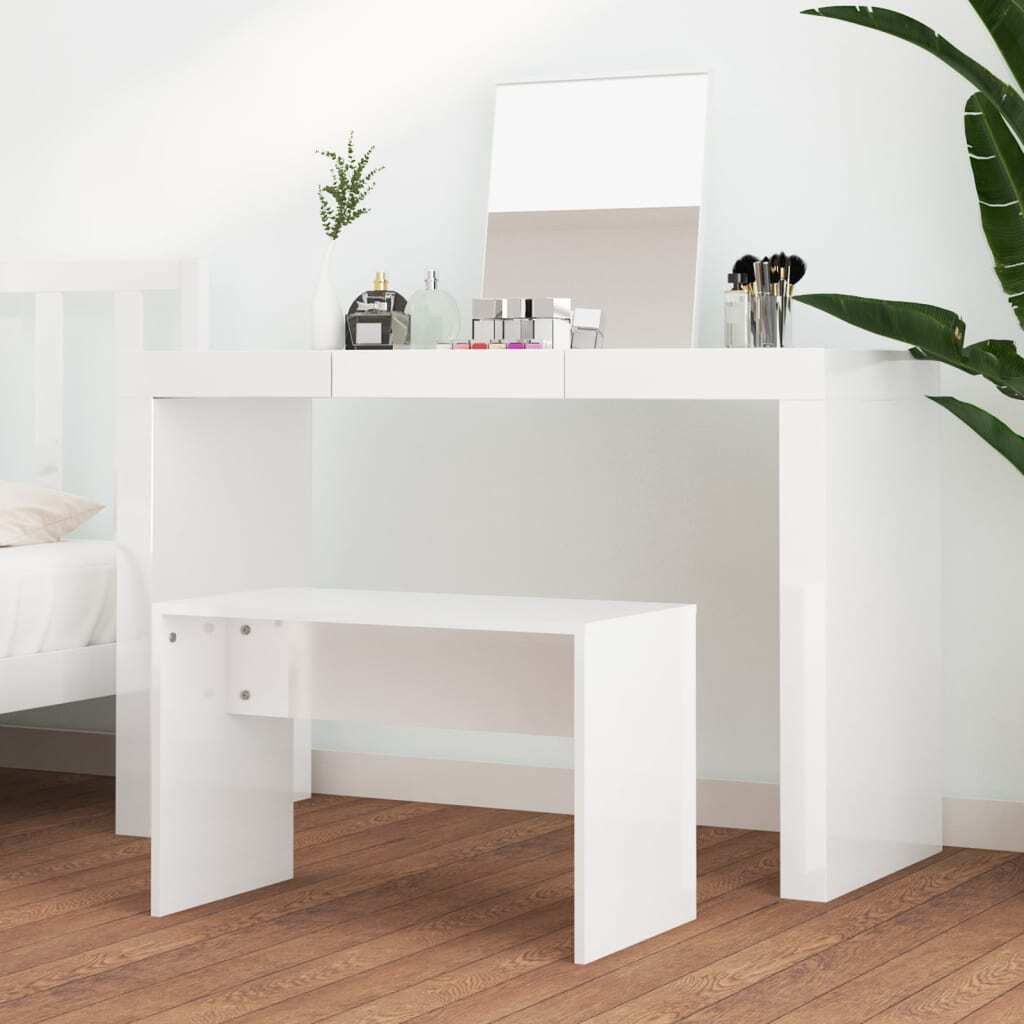 A white vanity table and stool set in a well-lit room, with a mirror and some makeup accessories on the table. A plant is visible to the right, and a bed is partially visible to the left.