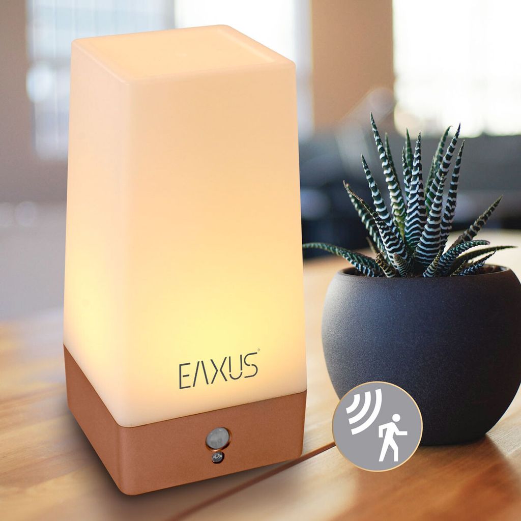 A bedside lamp with a motion sensor is placed on a wooden table next to a potted plant.