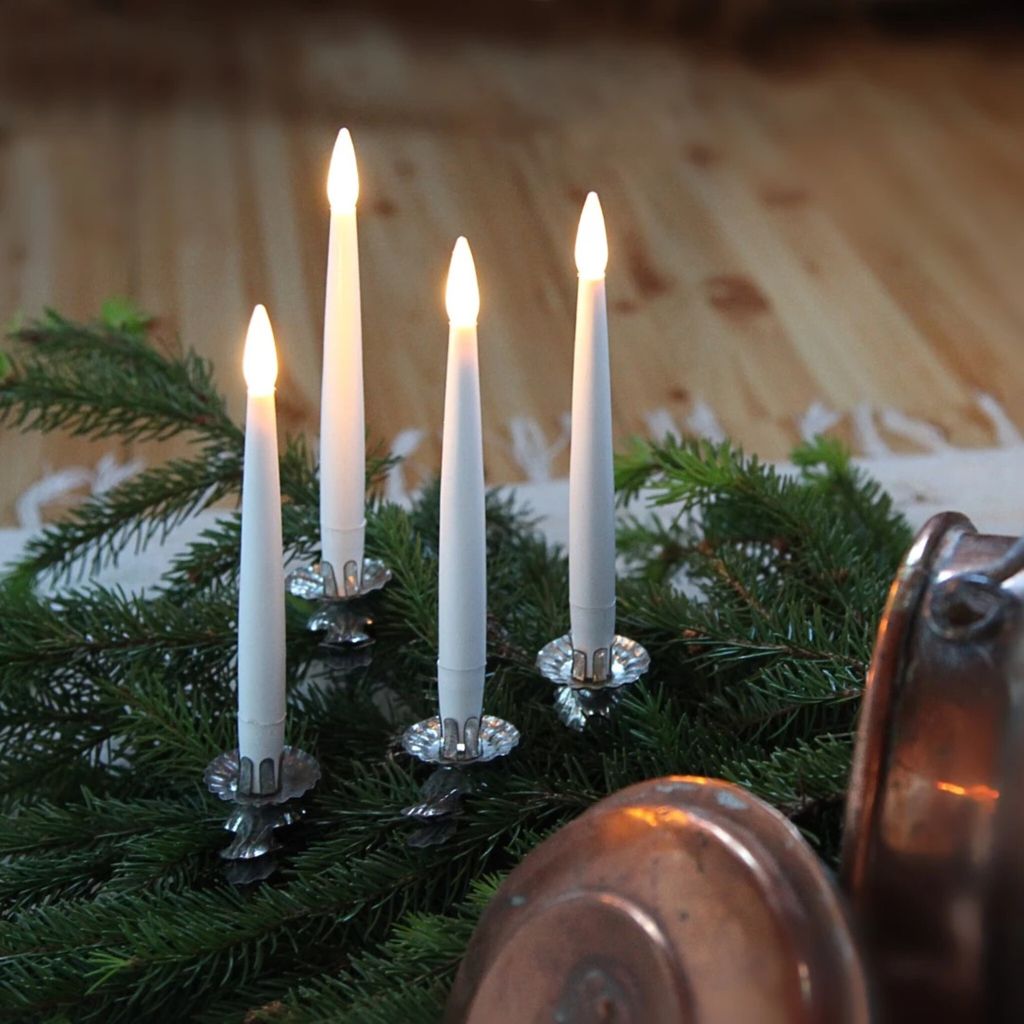 Four LED taper candles with flickering flames are arranged on a bed of evergreen branches, with copper pots in the foreground.