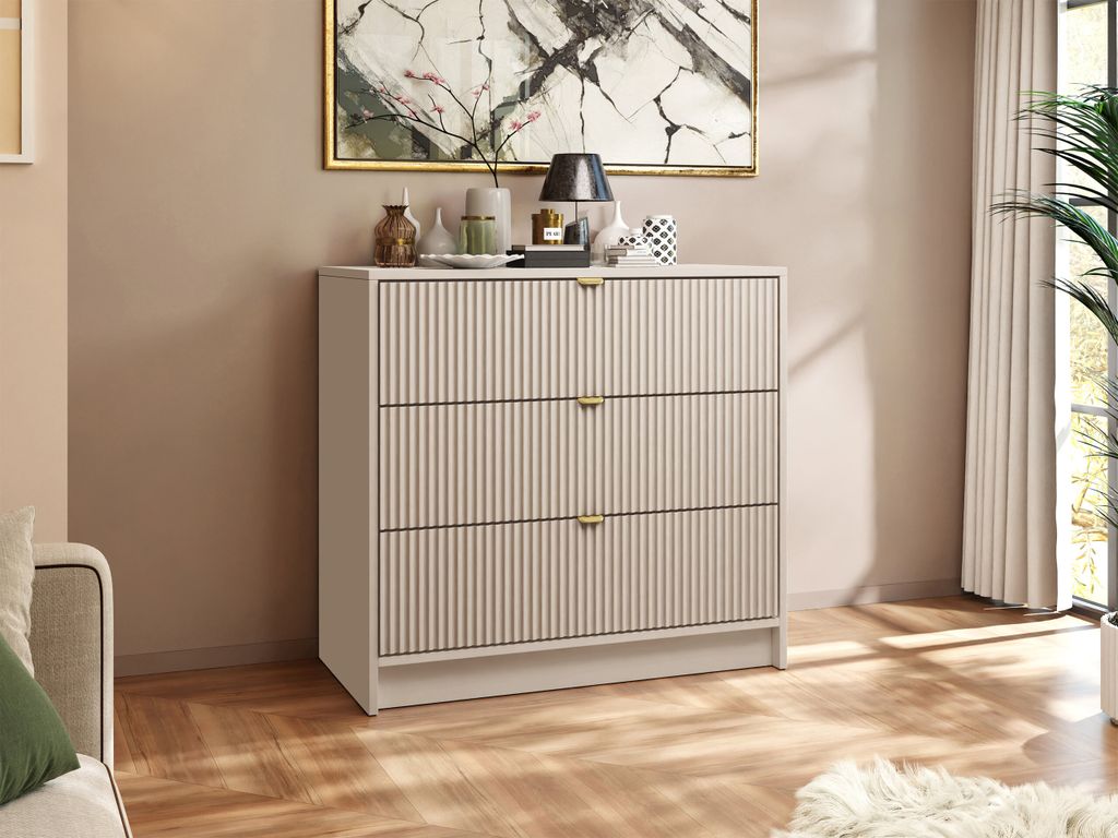 A chest of drawers with gold handles is placed in a room with a painting on the wall and a plant in the background.
