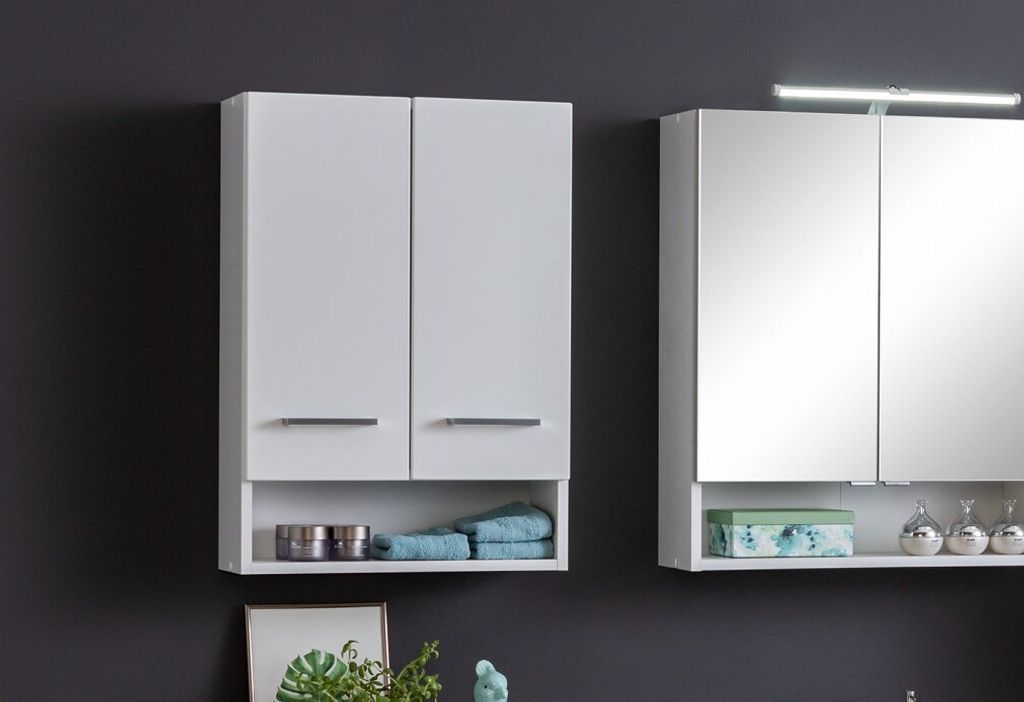 Two white bathroom cabinets are shown against a dark gray wall. The cabinet on the left has two doors and an open shelf below, containing towels and containers. The cabinet on the right has mirrored doors and an open shelf below, holding decorative items a