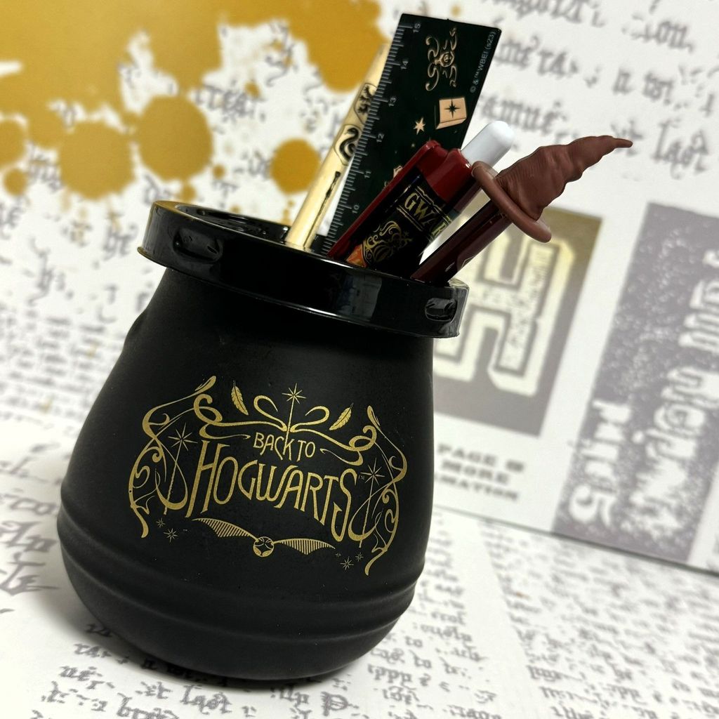 A close-up of a Harry Potter-themed cauldron pencil holder filled with pens and a ruler, set against a background of parchment-like paper with text and gold splatters.