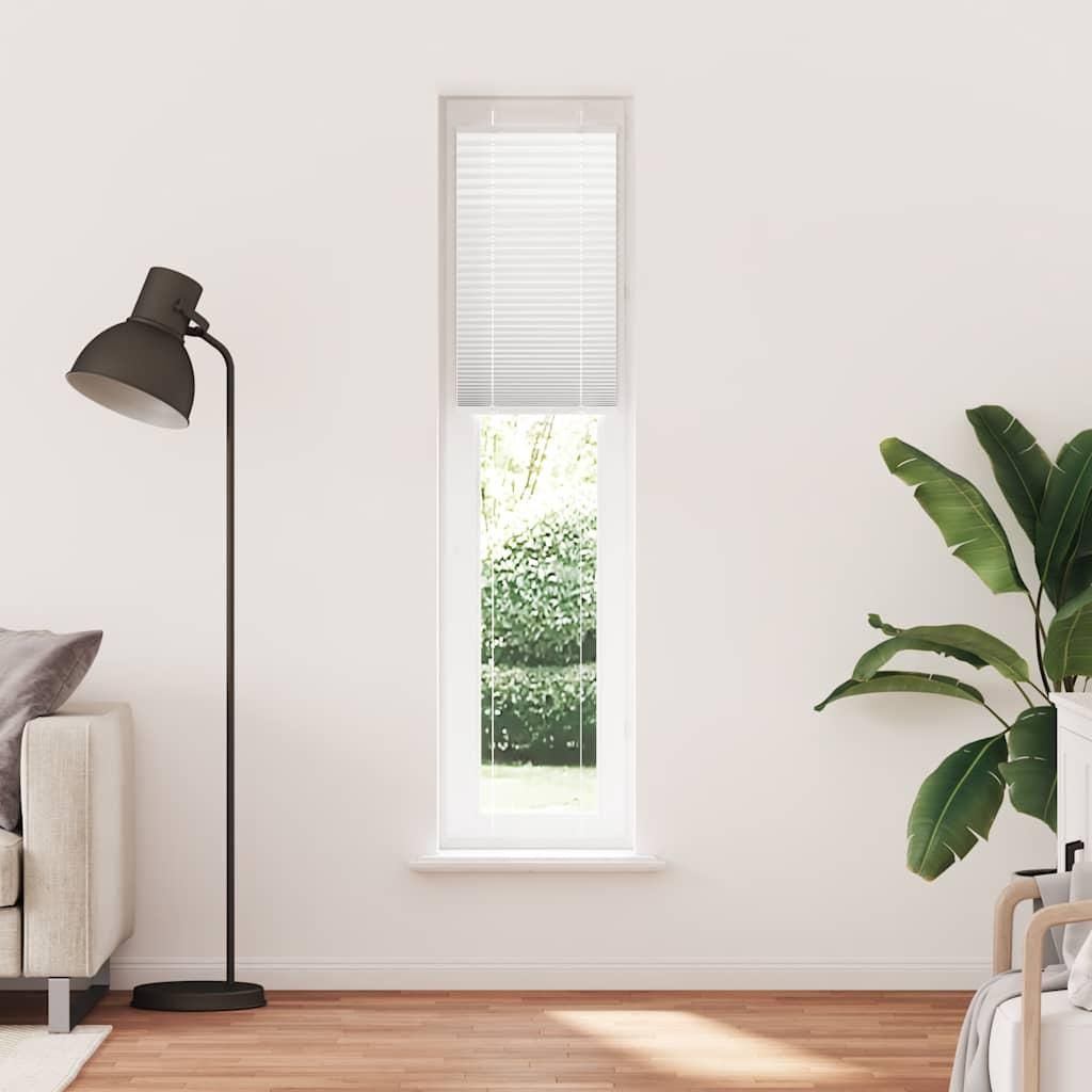 A tall, narrow window with white blinds is shown in the center of a white wall. To the left, a floor lamp and part of a sofa are visible. To the right, a large potted plant with broad green leaves is present. The floor is made of wooden planks.