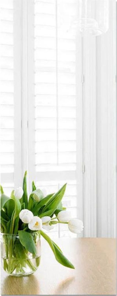 A vase of white tulips sits on a wooden table in front of white shutters.