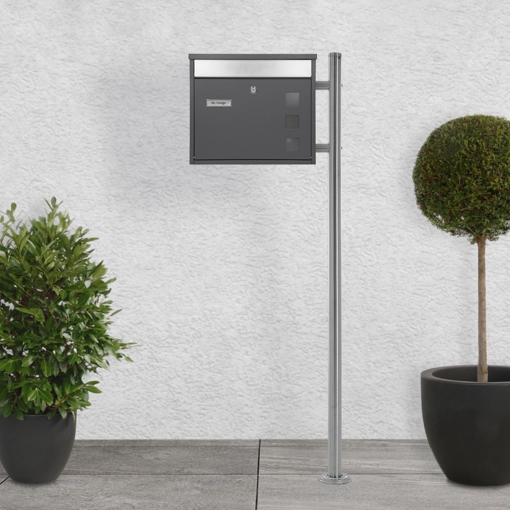 A mailbox with a newspaper compartment mounted on a stand, placed outdoors next to a wall and potted plants.