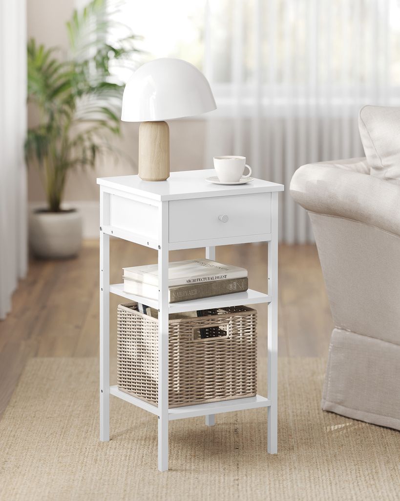 A white nightstand with a lamp, cup, books, and a basket on it, placed in a room with a plant and part of a couch visible.