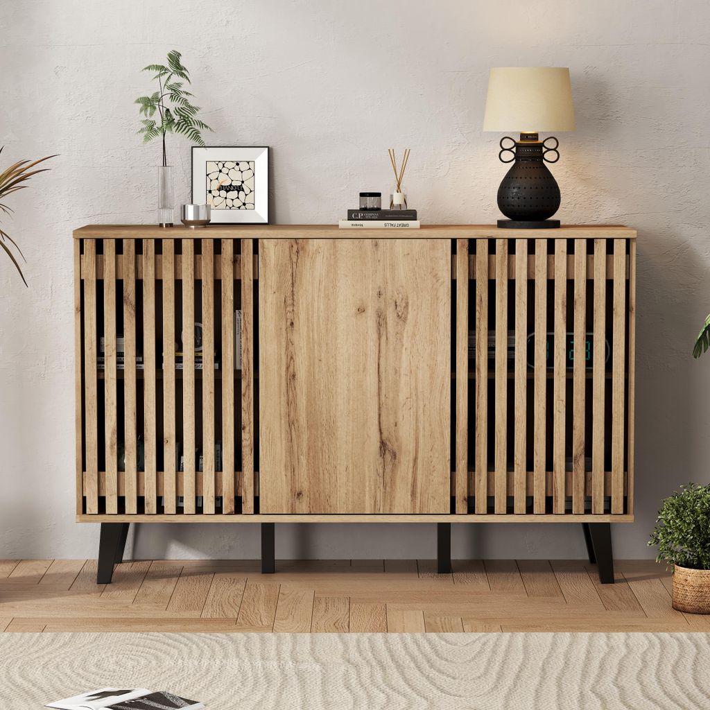 A wooden sideboard with slatted doors and black legs, decorated with a lamp, a framed picture, and a vase with a plant.