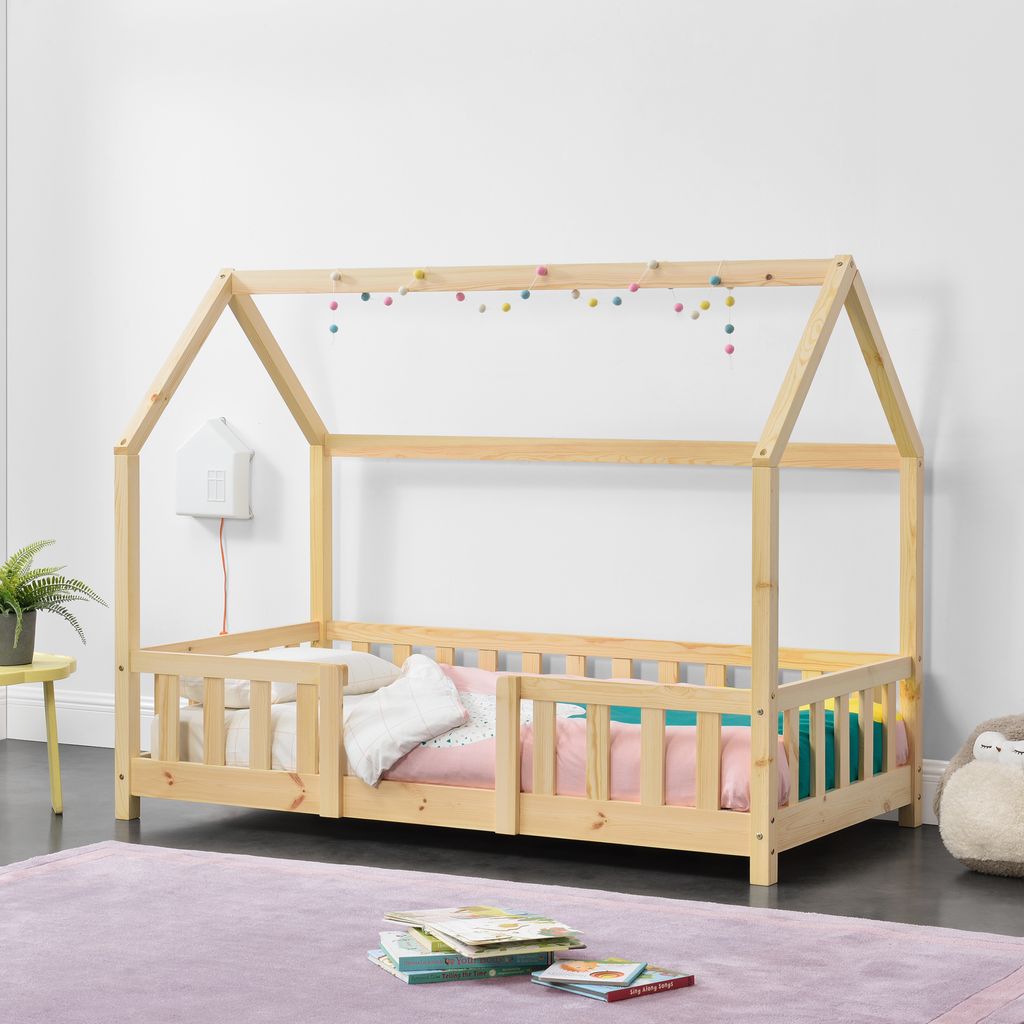 A child's house-shaped bed with wooden frame and railings, decorated with colorful pom-poms.
