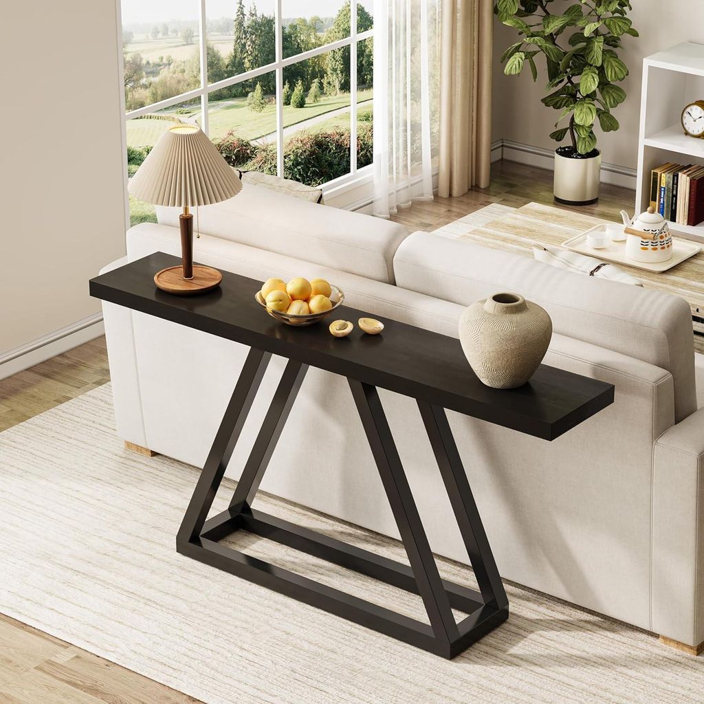 A dark wood console table with a modern geometric base is placed behind a white sofa in a well-lit living room. The table is decorated with a lamp, a bowl of yellow fruit, and a beige vase. A large window with a scenic view is in the background.