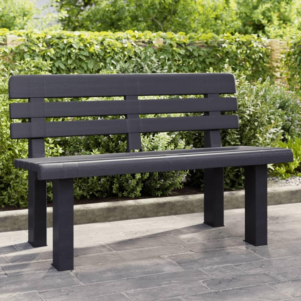 A dark gray garden bench is shown outdoors, with a hedge and paved ground in the background.