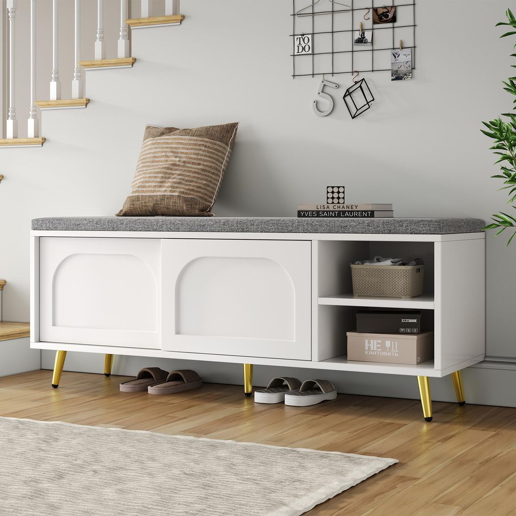 A white shoe bench with a gray cushion is placed in a hallway next to a staircase. There are shoes underneath the bench and decorative items on the wall above.