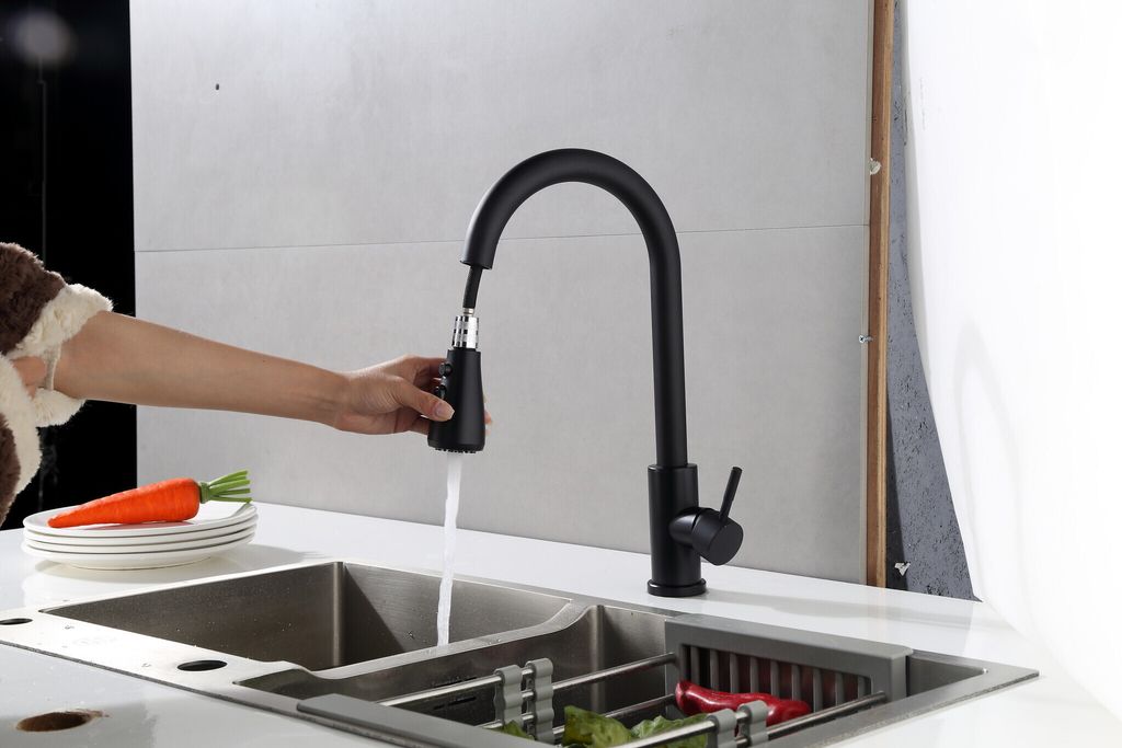 A person is using a black kitchen faucet with a pull-down sprayer over a double sink. There are plates and a carrot on the counter.