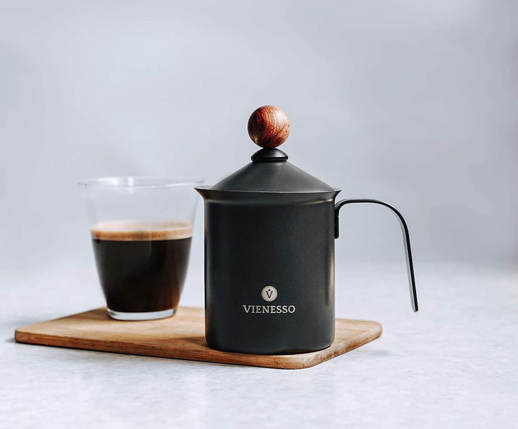 A black milk frother with a wooden knob on top sits next to a glass of coffee on a wooden board.