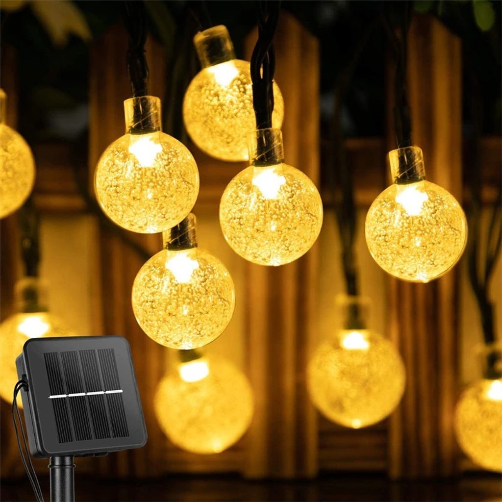 A string of solar-powered garden lights with round, textured bulbs, illuminated with warm white light, hanging in front of a wooden fence.
