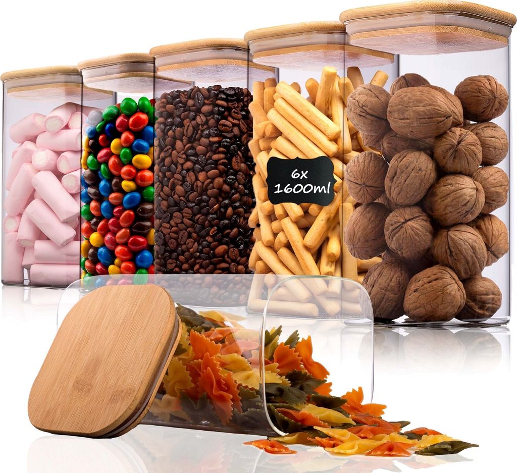 A collection of clear storage jars with bamboo lids, filled with various food items like marshmallows, candies, coffee beans, crackers, walnuts, and pasta. One jar has a chalk-like label indicating its capacity.
