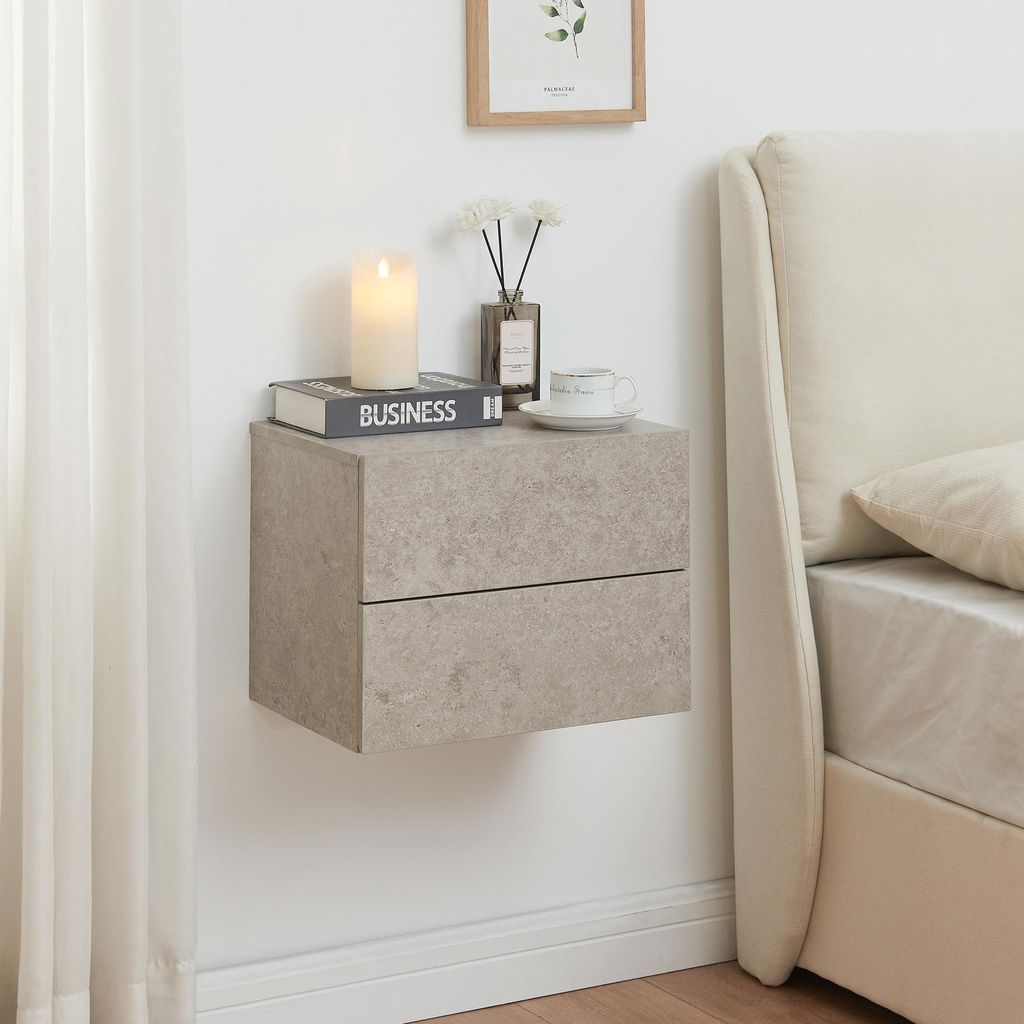 A floating nightstand with two drawers is mounted on a white wall next to a bed. The nightstand has a concrete-like texture and is decorated with a candle, a book, a vase with flowers, and a cup of coffee.