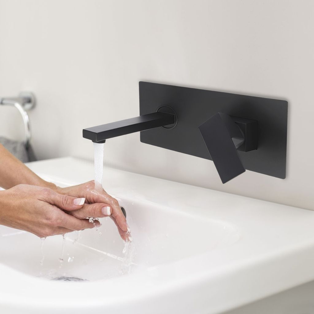 A person is washing their hands under a black wall-mounted faucet in a white sink.