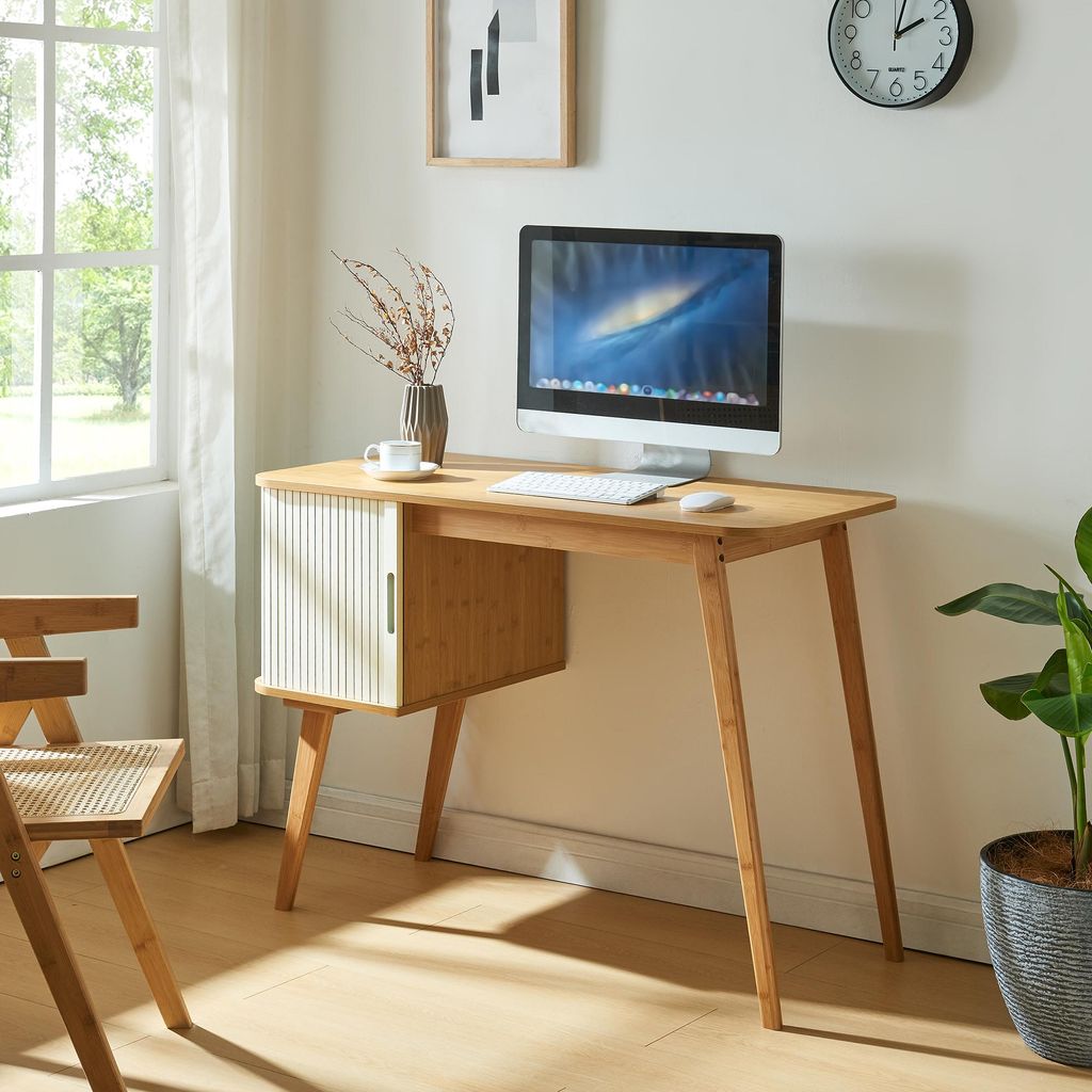 A desk with a computer and a plant in a room with a window.