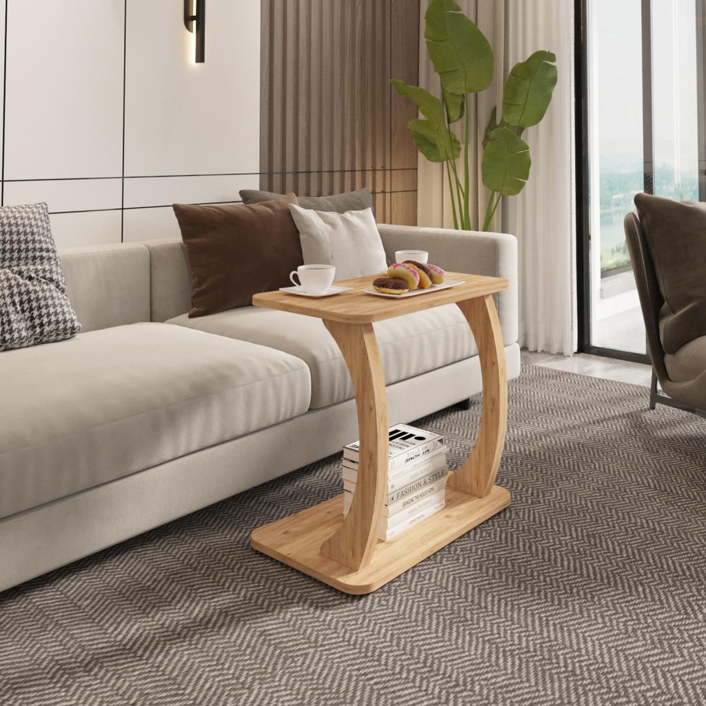 A wooden C-shaped side table is placed next to a cream-colored sofa in a living room. The table has a shelf with books and holds two cups and pastries.