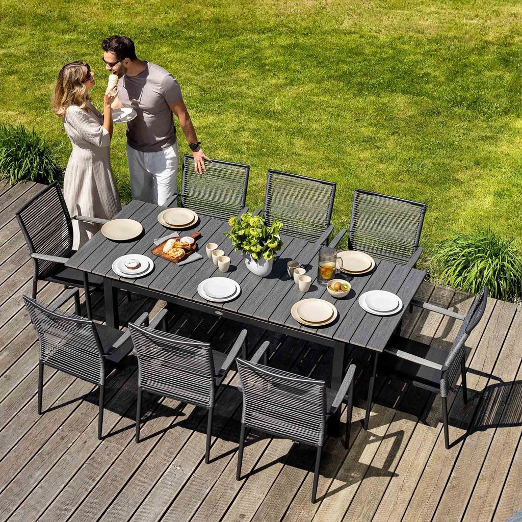 A couple is enjoying a meal at a dining table set up on a wooden deck in a garden.