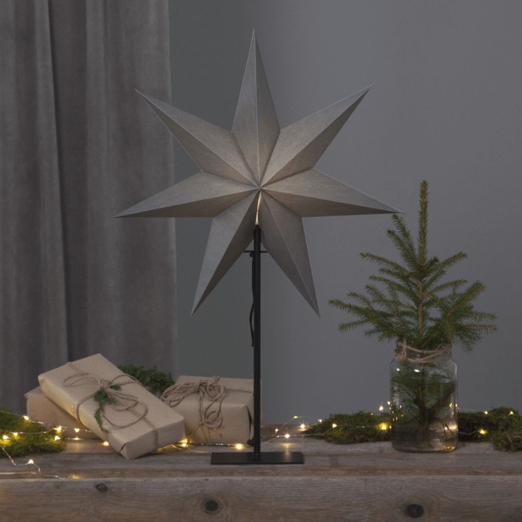 A large, grey, star-shaped lamp is displayed on a wooden surface, surrounded by wrapped gifts and small fairy lights. A miniature Christmas tree in a glass jar is also present, creating a festive, indoor holiday scene.