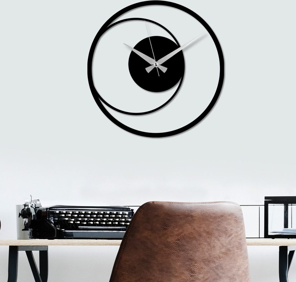 A modern black metal wall clock with a circular design is mounted on a white wall above a desk with a vintage typewriter.