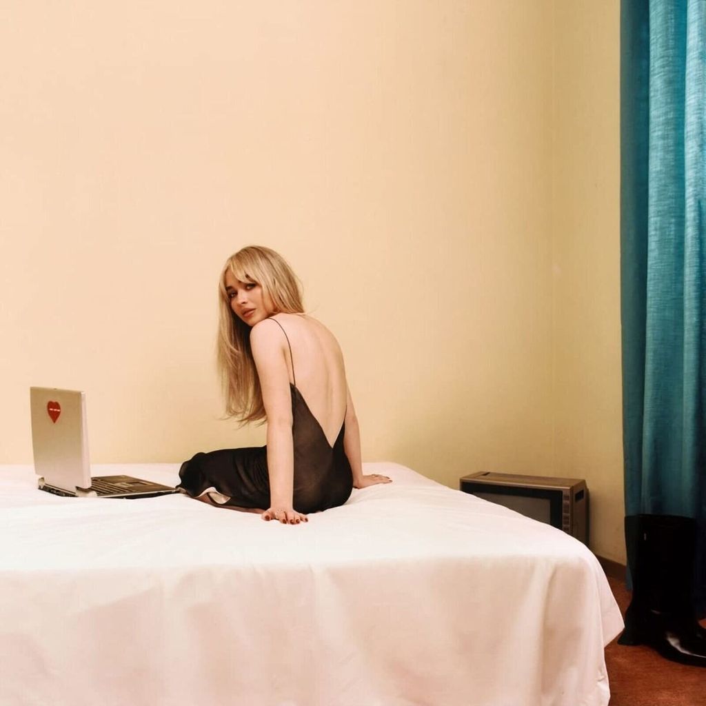 A woman sits on a bed next to a laptop, looking over her shoulder.