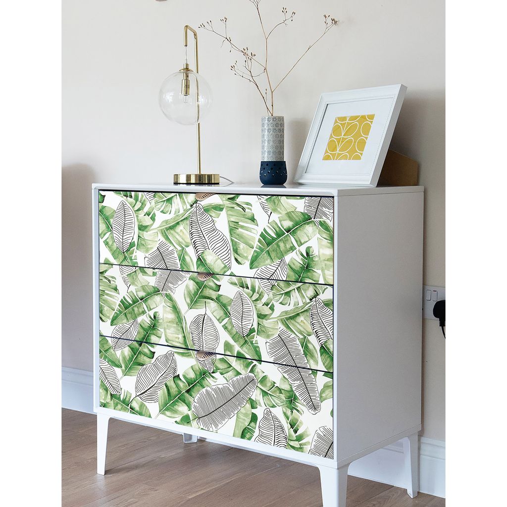 A white dresser with drawers decorated with a tropical leaf pattern, a gold lamp, and a framed picture on top.