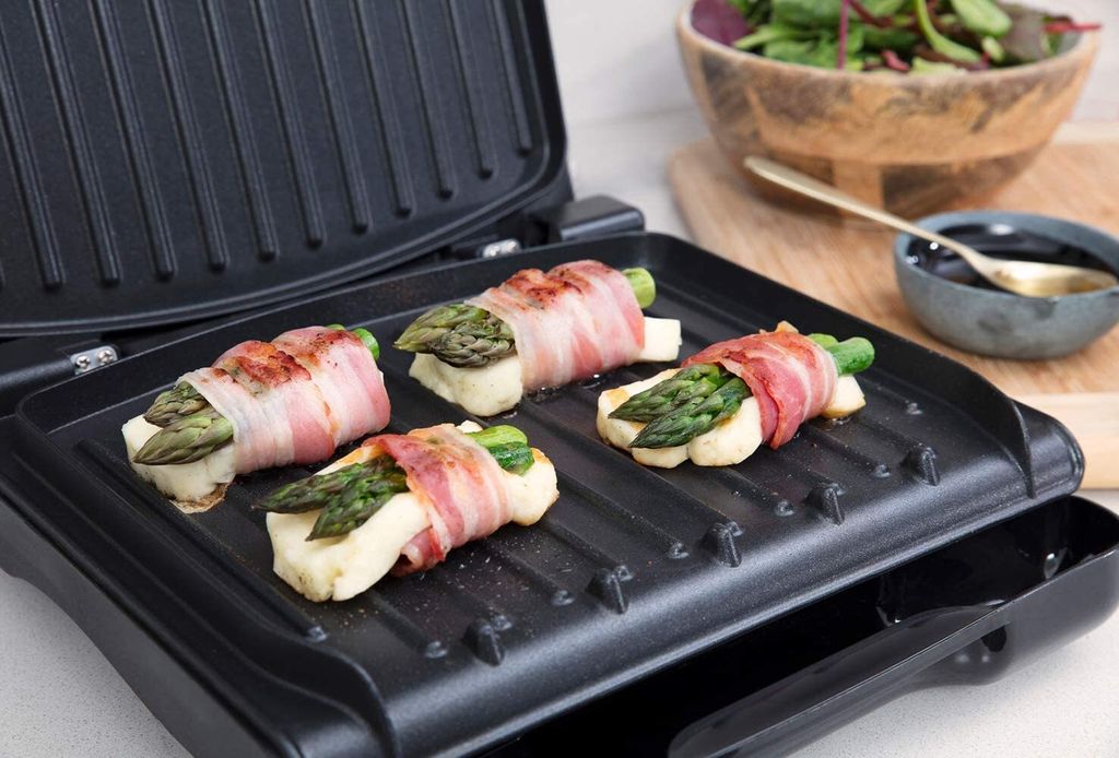 Asparagus and cheese wrapped in bacon are being grilled on a George Foreman grill. A salad and a bowl of sauce are visible in the background.