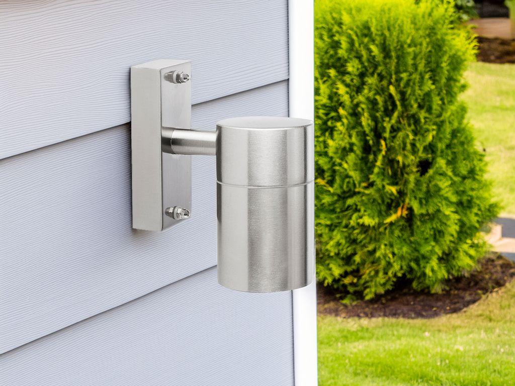 A silver outdoor wall light mounted on a light gray, horizontally sided wall, with a green bush and grass in the background.