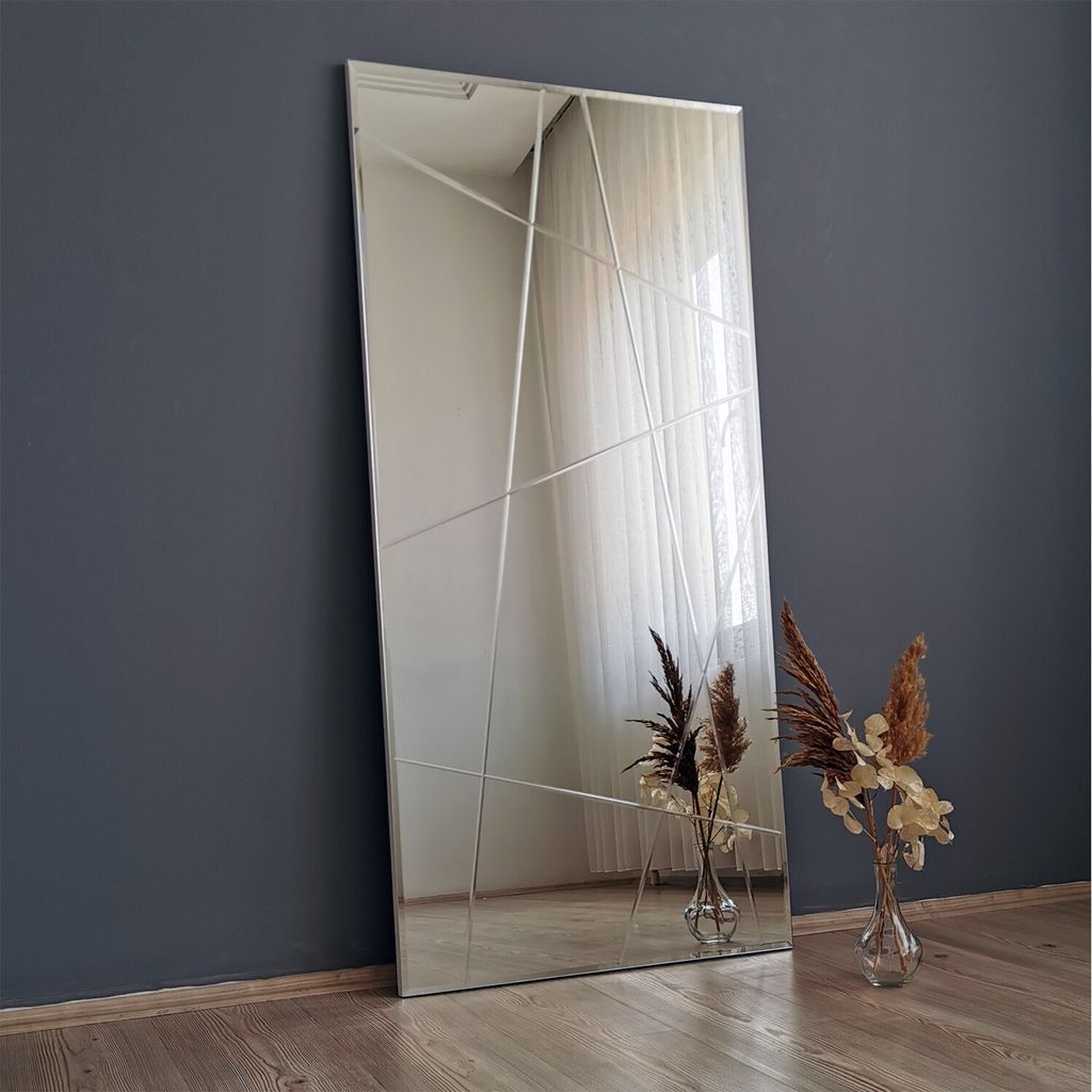 A full-length mirror with a geometric design, leaning against a dark gray wall. Two vases with dried flowers are placed on the floor in front of the mirror.