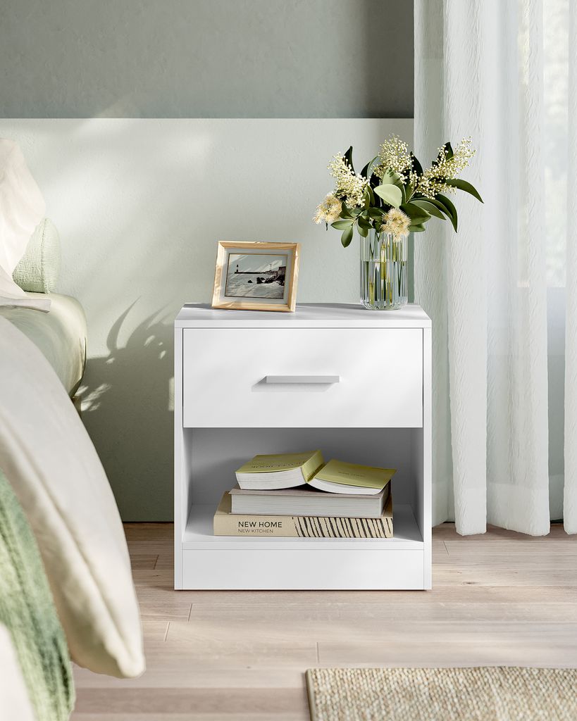 A white nightstand with a drawer and an open shelf, placed next to a bed. It holds a framed picture and a vase of flowers on top. Books are stacked in the open shelf.