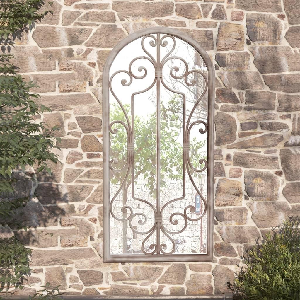 A decorative mirror with an ornate metal frame is mounted on a rustic stone wall, with greenery visible in the reflection and to the sides.